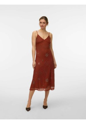 Vrouw die een mouwloze midi-jurk in roestkleur draagt met dunne bandjes, versierd met subtiele decoratieve patronen, staand tegen een effen achtergrond.