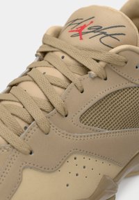 Gros plan d'une basket beige avec des lacets tissés, un panneau latéral en mesh, des détails perforés et un logo stylisé avec une figure rouge sur la languette.