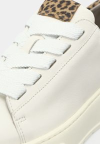 Baskets blanches avec de larges lacets texturés et un détail imprimé léopard sur la languette, montrant les coutures et la texture lisse du cuir.