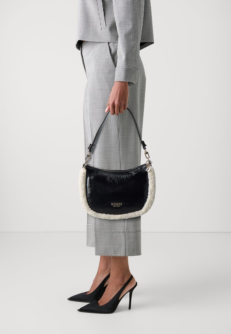 Personne en costume à carreaux noir et blanc tenant un sac à main noir GUESS avec une bordure en fourrure blanche, portant des chaussures slingback à talons hauts noires.