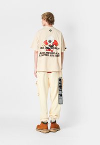 Camiseta de gran tamaño beige con gráfico en la parte posterior, pantalones cargo crema con bolsillo lateral y etiqueta negra, y botas marrones con cordones.