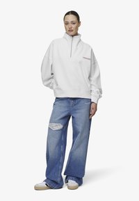 Sudadera de forro polar blanca con media cremallera, que presenta un logo en el pecho. Combinada con jeans de pierna ancha rasgados de color azul y zapatillas beige.