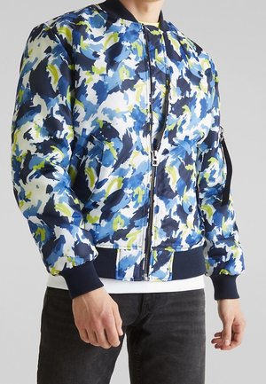 Mann trägt eine Bomberjacke mit abstraktem Muster in Blau, Weiß und Grün, mit Frontreißverschluss sowie gerippten Bündchen und Saum.