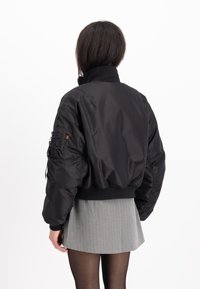Chaqueta bomber negra recortada con un acabado brillante, cuello y puños acanalados, y bolsillos laterales. Presenta un bolsillo en la manga detallado con cremallera.
