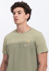 Olive green cotton t-shirt with a round neckline features a front illustration of a child near a structure with flames and a mountain backdrop.