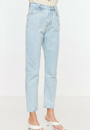 Vrouw met lichtblauwe jeans met hoge taille en een losse pasvorm, en witte open-teen sandalen met hak op een effen achtergrond.