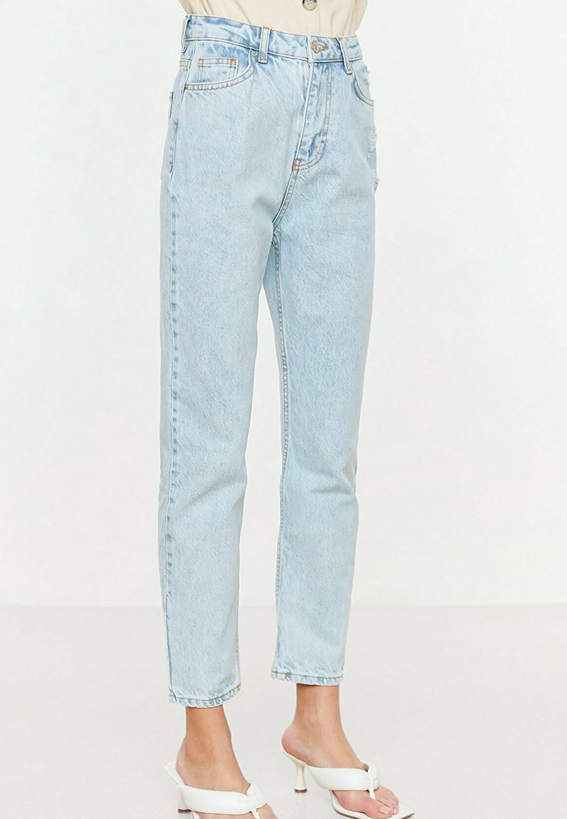 Femme portant un jean bleu clair taille haute à coupe décontractée et des sandales blanches ouvertes à talons sur un fond uni.
