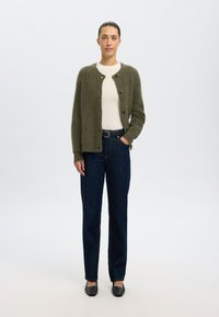 Olivgrön stickad kappa med rund hals och knäppning, bärs över en krämfärgad topp och mörkblå raka jeans, kombinerat med svarta lägenheter.