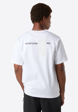 Mand iført hvid t-shirt med teksten "NEVER STOP EXPLORING" på ryggen, kombineret med sorte bukser, står foran en ensfarvet hvid baggrund.