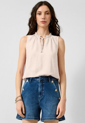 Femme portant un haut sans manches beige clair à col volanté et lien, associé à un short en denim bleu taille haute avec des boutons dorés.