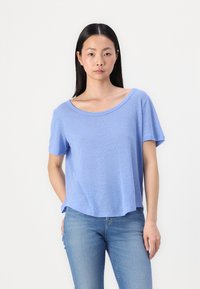 Tricou de culoare albastru deschis, cu mânecă scurtă, gât larg, textură moale și croială lejeră. Asortat cu blugi albăstrii pentru un aspect casual.