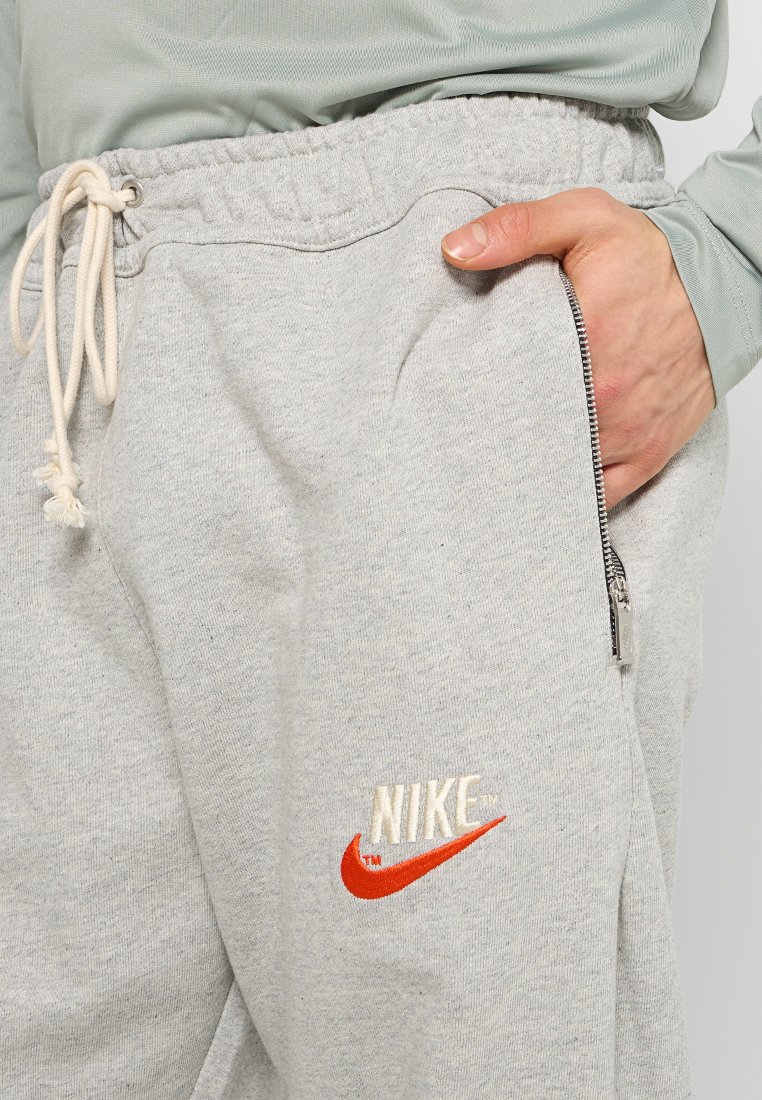 Pantalon de survêtement gris avec une taille élastique, un cordon de serrage, une poche latérale zippée et un logo Nike brodé en orange et blanc. Tissu doux et texturé.