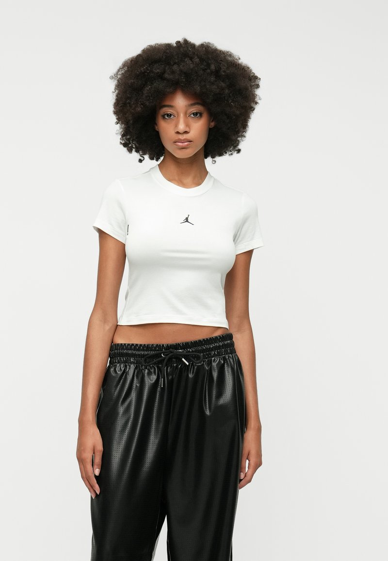 Λευκό κοντό cropped t-shirt με κοντά μανίκια, με μικρό μαύρο λογότυπο στο στήθος, φορεμένο με μαύρο παντελόνι φόρμας με κορδόνι στη μέση.