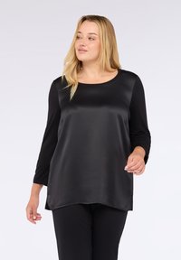 Donna bionda che indossa un top in satin nero a maniche lunghe e pantaloni neri, che cammina con un'espressione neutra su uno sfondo semplice.