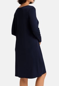 Donna che indossa un abito blu navy di lunghezza al ginocchio e maniche lunghe, vista da dietro con le mani posizionate vicino ai fianchi.