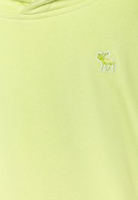 Svetlá žltá mikina s kapucňou s malým zeleným vyšívaným logom losa vpredu. Vyrobená z mäkkej tkaniny s kapucňou na šnurovanie.