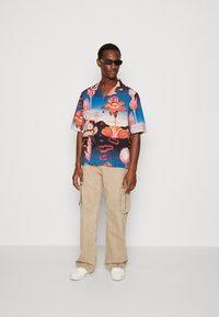 Cămașă cu mânecă scurtă de un albastru vibrant, cu modele florale; asortată cu pantaloni cargo bej deschis și adidași albi. Ochelari de soare rectangulari negri.