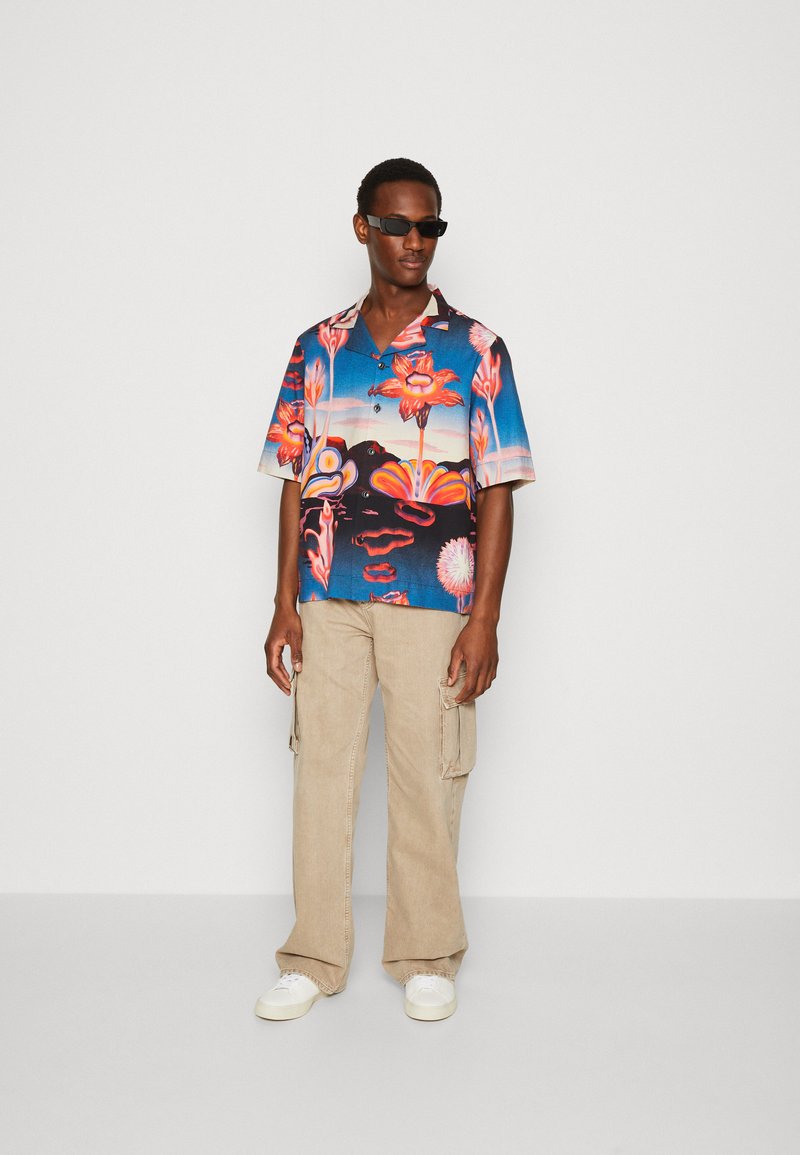 Cămașă cu mânecă scurtă de un albastru vibrant, cu modele florale; asortată cu pantaloni cargo bej deschis și adidași albi. Ochelari de soare rectangulari negri.