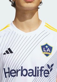 Vit idrottströja med svarta diagonala ränder, gula axelinslag, adidas-logotyp, LA Galaxy-sköld och "Herbalife"-branding i marinblå.