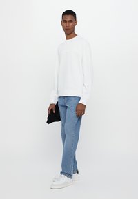 White sweatshirt with embroidered text, paired with light blue denim jeans and white sneakers. Model holding a black item.