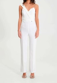 Combinaison blanche au design ajusté, avec une bretelle sur une épaule et un accent chaîne. Présente un décolleté en V profond et des jambes larges. Texture lisse.