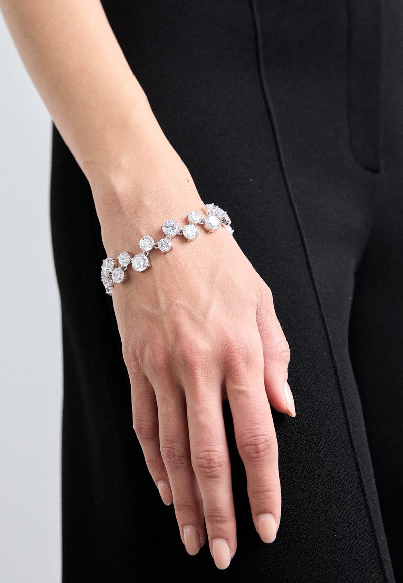 Ασημένιο βραχιόλι με εναλλασσόμενες στρογγυλές και τετράγωνες διαφανείς ρόμβους, σε λεπτό σχέδιο, φορεμένο σε χέρι με φόντο μαύρο.