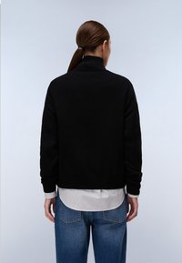 Mustat neulepusero, jossa on korkea kaulus, ribattu helma ja pitkät hihat, kerrostettuna valkoisen ja harmaan raidallisen paidan päälle, yhdistettynä sinisiin farkkuihin.