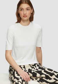 Pull blanc à manches courtes avec un col rond, texture lisse et coupe ajustée. Le modèle le porte avec un pantalon noir et beige à motifs.