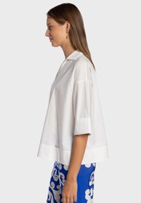 Blouse blanche à manches courtes avec col, présentant une coupe décontractée et un subtil accent métallique à l'ourlet. Texture lisse, tissu léger.