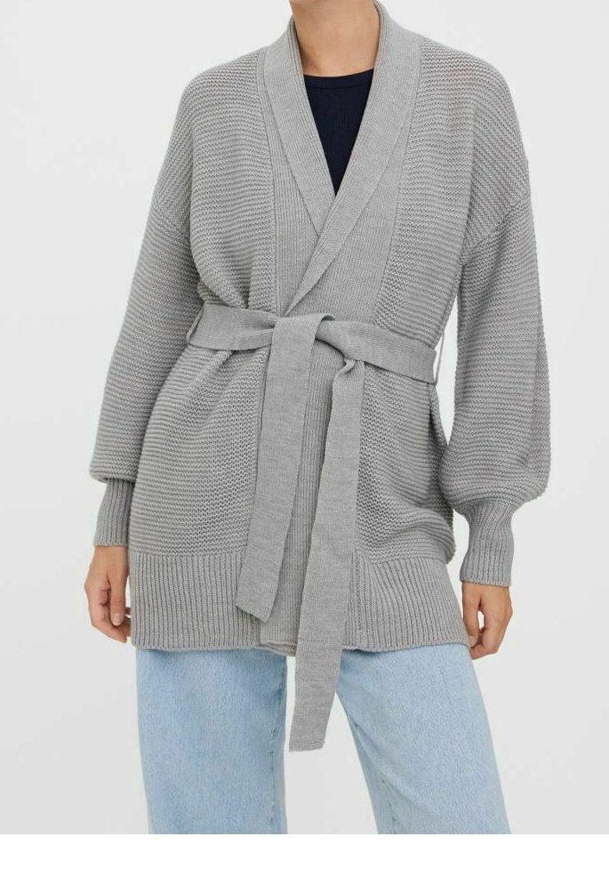Femme portant un cardigan en maille gris clair avec une ceinture nouée à la taille, associé à un jean bleu clair et une chemise foncée en-dessous.