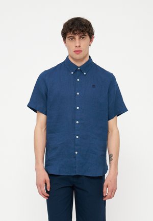 Giovane uomo con capelli castani ricci che indossa una camicia blu scuro a maniche corte con bottoni e pantaloni abbinati, in piedi davanti a uno sfondo bianco semplice.