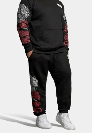 Homme portant un sweat-shirt noir et un pantalon avec des motifs de dragon rouges et blancs sur les manches et la jambe, assortis à des baskets blanches.