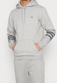 Šedá mikina s kapucí, přední kapsou, nastavitelnou kapucí a pruhovanými šedými detaily na rukávech. Obsahuje malé logo na hrudi.