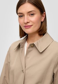 Beige trenchcoat met een grote kraag, twee knopen aan de voorkant en een gladde textuur, gedragen over een wit shirt. Eenvoudig, klassiek ontwerp.