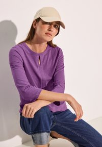Lila Langarmshirt mit rundem Ausschnitt und Schlüssel-Eingangsdetail, kombiniert mit blauen, hochgekrempelten Jeans und einer beigen Cap.