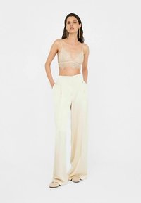 Beige lace bralette and cream wide-leg pants, featuring a smooth texture, high waist, and side pockets. Model stands with hands in pockets.