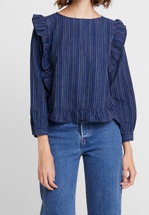 Vrouw draagt een marineblauwe blouse met verticale strepen en ruches op mouwen en zoom, gecombineerd met een medium gewassen blauwe spijkerbroek.
