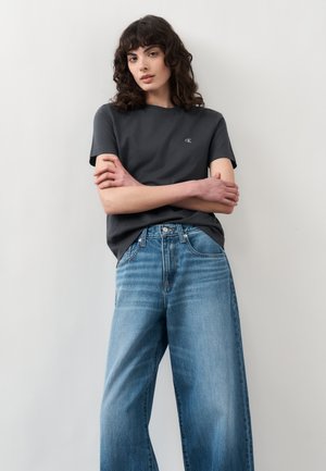 Νεαρή γυναίκα με σγουρά σκούρα μαλλιά φοράει μαύρο κοντομάνικο μπλουζάκι και φαρδιά μπλε τζιν, στέκεται με σταυρωμένα χέρια μπροστά σε ένα απλό φόντο.