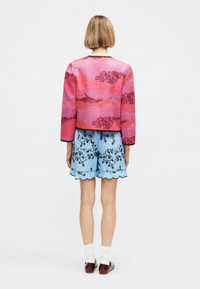 Γυναίκα με κοντά ξανθά μαλλιά που φοράει ροζ και κόκκινο μοτίβο στο μπουφάν, ανοιχτό μπλε φλοράλ σορτς με κυματιστές άκρες, λευκές κάλτσες και καφέ παπούτσια, με την πλάτη γυρισμένη.