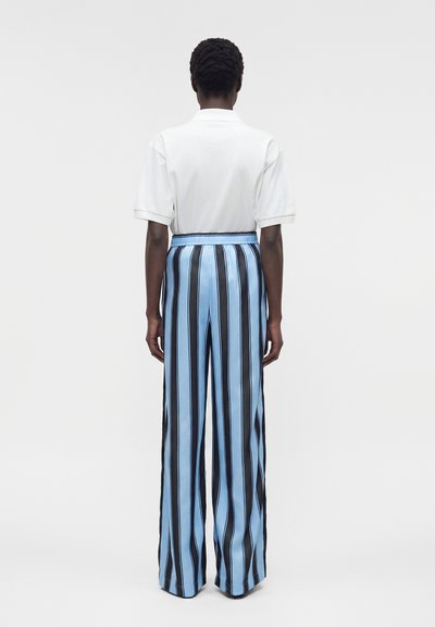 Άτομο που στέκεται με την πλάτη γυρισμένη, φοράει λευκό κοντομάνικο πουκάμισο και ψηλόμεσο, φαρδύ παντελόνι με κάθετες μπλε και μαύρες ρίγες.