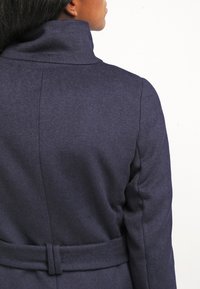 Personne portant un manteau en laine bleu foncé avec un col haut et une ceinture, vue de dos et légèrement tournée sur le côté.