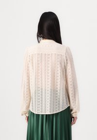 Lace blouse in off-white with long sleeves, intricate patterns, rounded hem, and a delicate collar, worn over a pleated green skirt.
