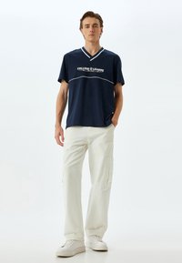 Marineblå bomulds t-shirt med hvid tekst "COLLEGE LEAGUE", V-hals design, parret med kremfarvede cargo bukser og hvide sneakers.