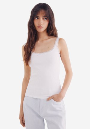 White tank top with a lace trim, made of ribbed fabric, featuring thin shoulder straps and a fitted shape. Paired with striped pajama pants.
