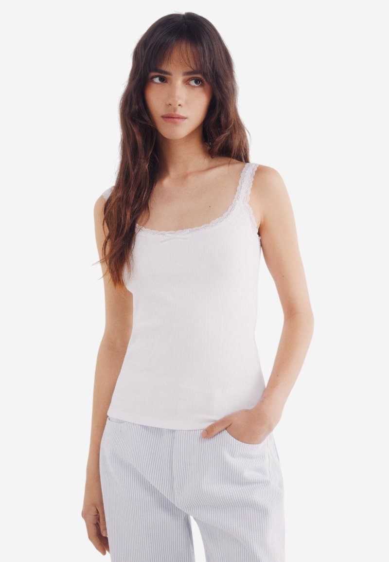 Top bianco senza maniche con bordo in pizzo, realizzato in tessuto a coste, con spalline sottili e forma aderente. Abbinato a pantaloni da pigiama a righe.