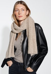 Beige sjaal met structuur om de nek van een model gewikkeld, gecombineerd met een zwarte leren jas; het model heeft lang haar en een neutrale top.