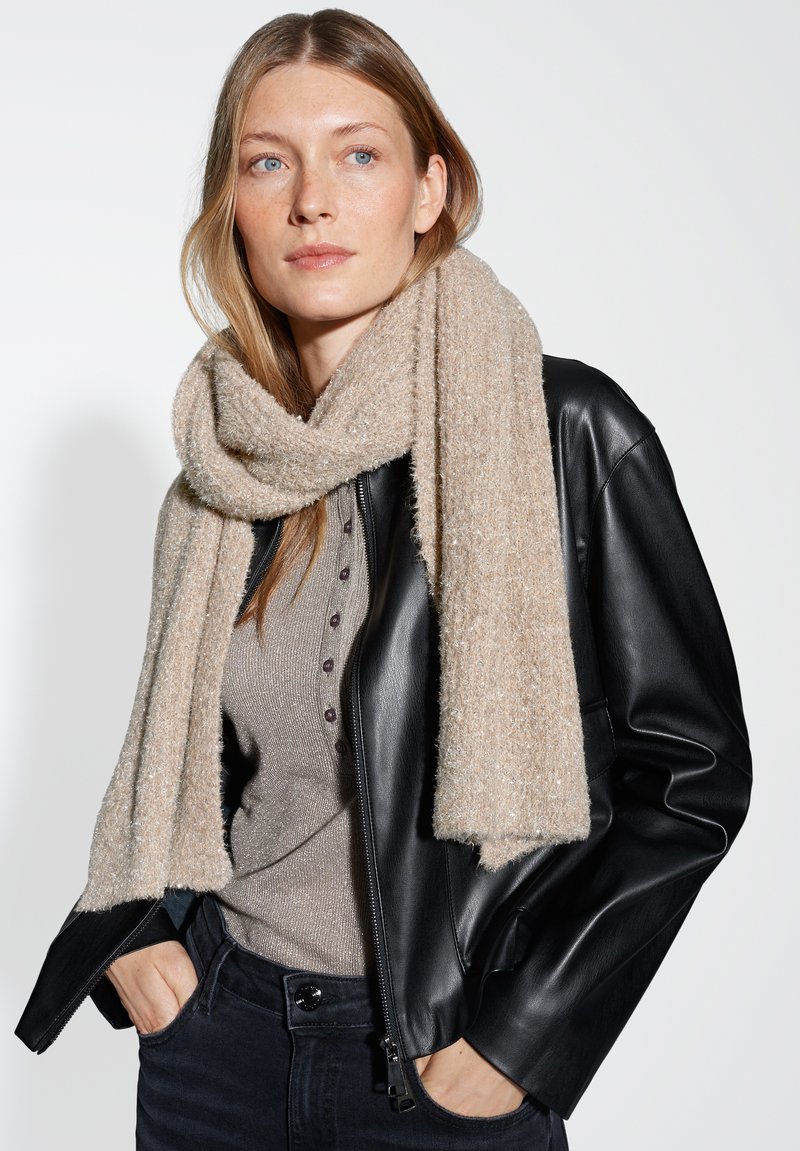 Beige sjaal met structuur om de nek van een model gewikkeld, gecombineerd met een zwarte leren jas; het model heeft lang haar en een neutrale top.