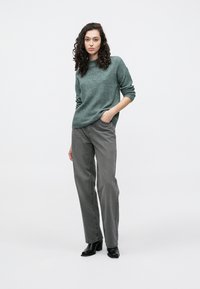 Strickpullover in Blaugrün, graue hoch taillierte Jeans und schwarze Stiefel mit Absatz. Das Modell steht mit entspannten Armen, der Hintergrund ist schlicht weiß.