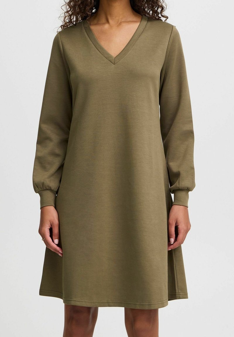 Femme portant une robe longue à manches longues de couleur vert olive avec un décolleté en V et des manches retroussées, debout devant un fond clair uni.