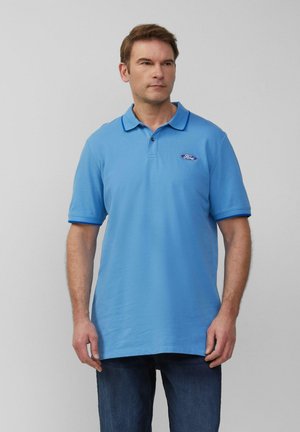 Man in een lichtblauw polo-shirt met Ford-log en donkere spijkerbroek, staand tegen een effen lichtgrijze achtergrond.